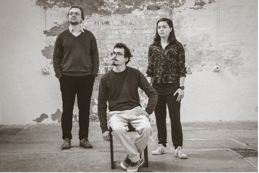 Eine Schwarz-Weiß-Fotografie der drei Musiker:innen der Band "Die Schlaue Bildkröte" vor einer weißen Wand. Alle drei Personen schauen nach links oben in die Ferne.