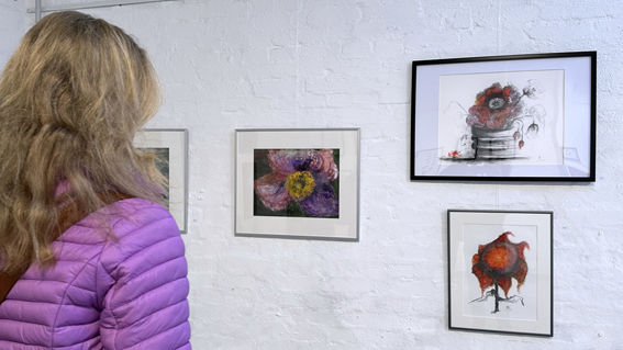Eine Person schaut sich gerahmte Kunstwerke an. Die Fotografie zeigt die Person von hinten und vier Zeichnungen, die Blumen darstellen.